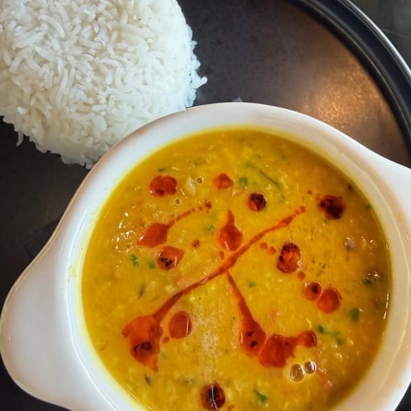 A white bowl of yellow lentil soup with red oil swirls, next to a mound of white rice.