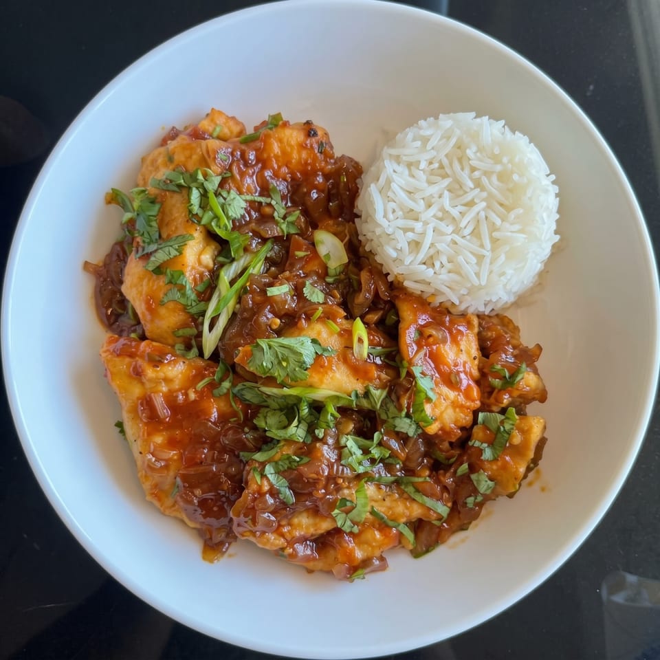 A white bowl filled with chicken in a rich brown sauce, garnished with cilantro and green onions, served with white rice.