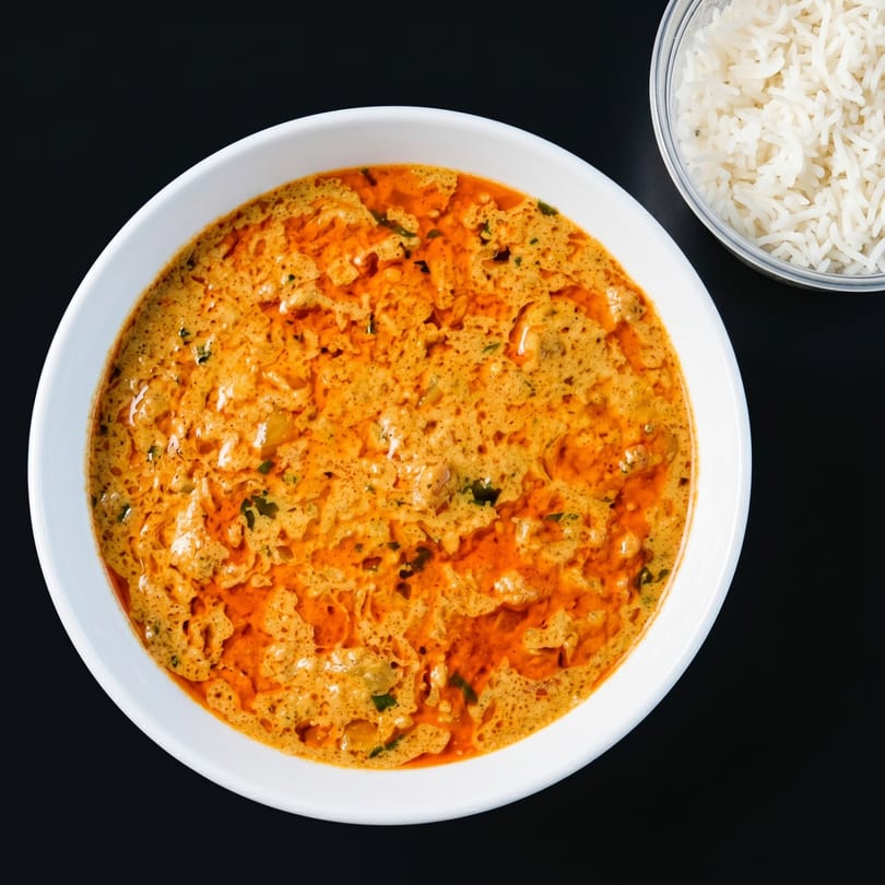 A white bowl of creamy orange curry with green herbs, next to a container of white rice.