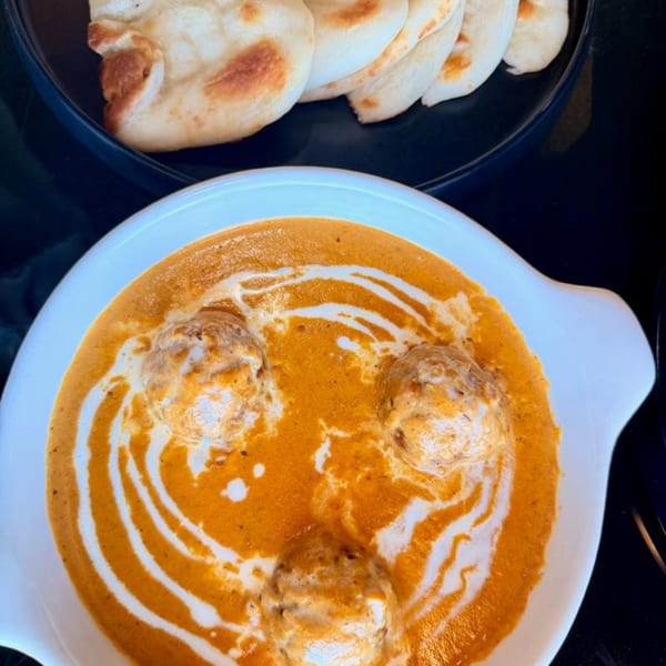 A white bowl of creamy orange curry with three meatballs and swirled cream, served with naan bread.