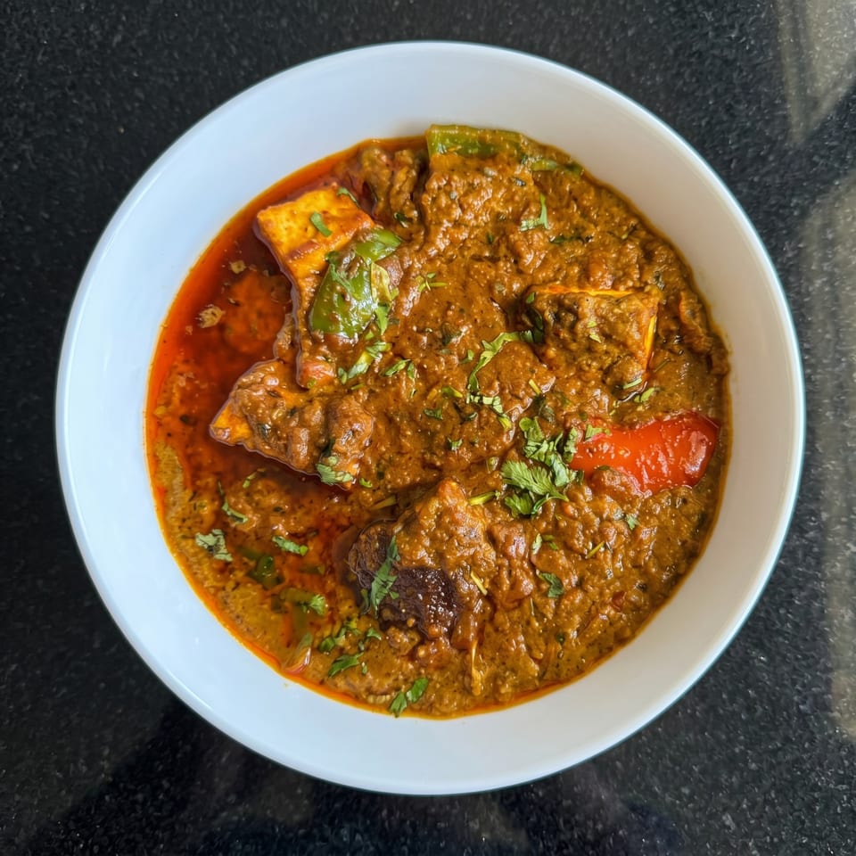 A white bowl filled with a rich, reddish-brown curry, garnished with green herbs and red peppers.